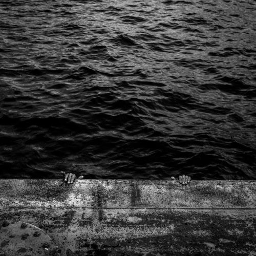 Fotografia horizontal em preto e branco com imagemdas águas escuras do rio Negro com reflexos de luzes acima e na parte de baixo da imagem, parte de um muro de concreto com duas mão que se apoiam